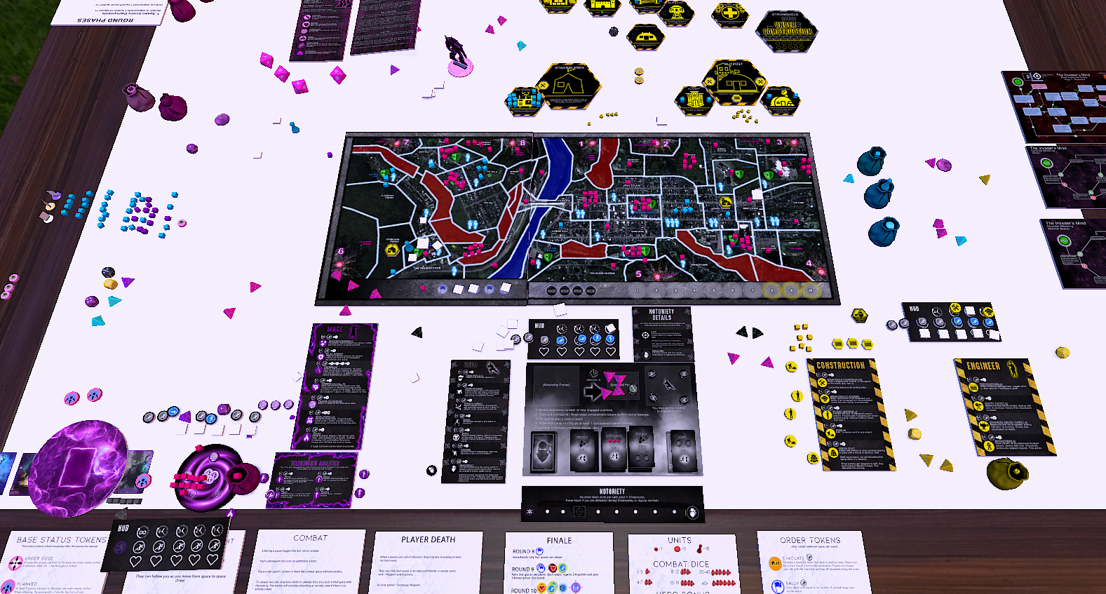 An overhead view of the JT Free board game laid out on a virtual tabletop, showing a city map board with red and blue territory lines, dozens of resource tokens in pink and yellow, character cards along the edges, and a row of player ability cards across the bottom.